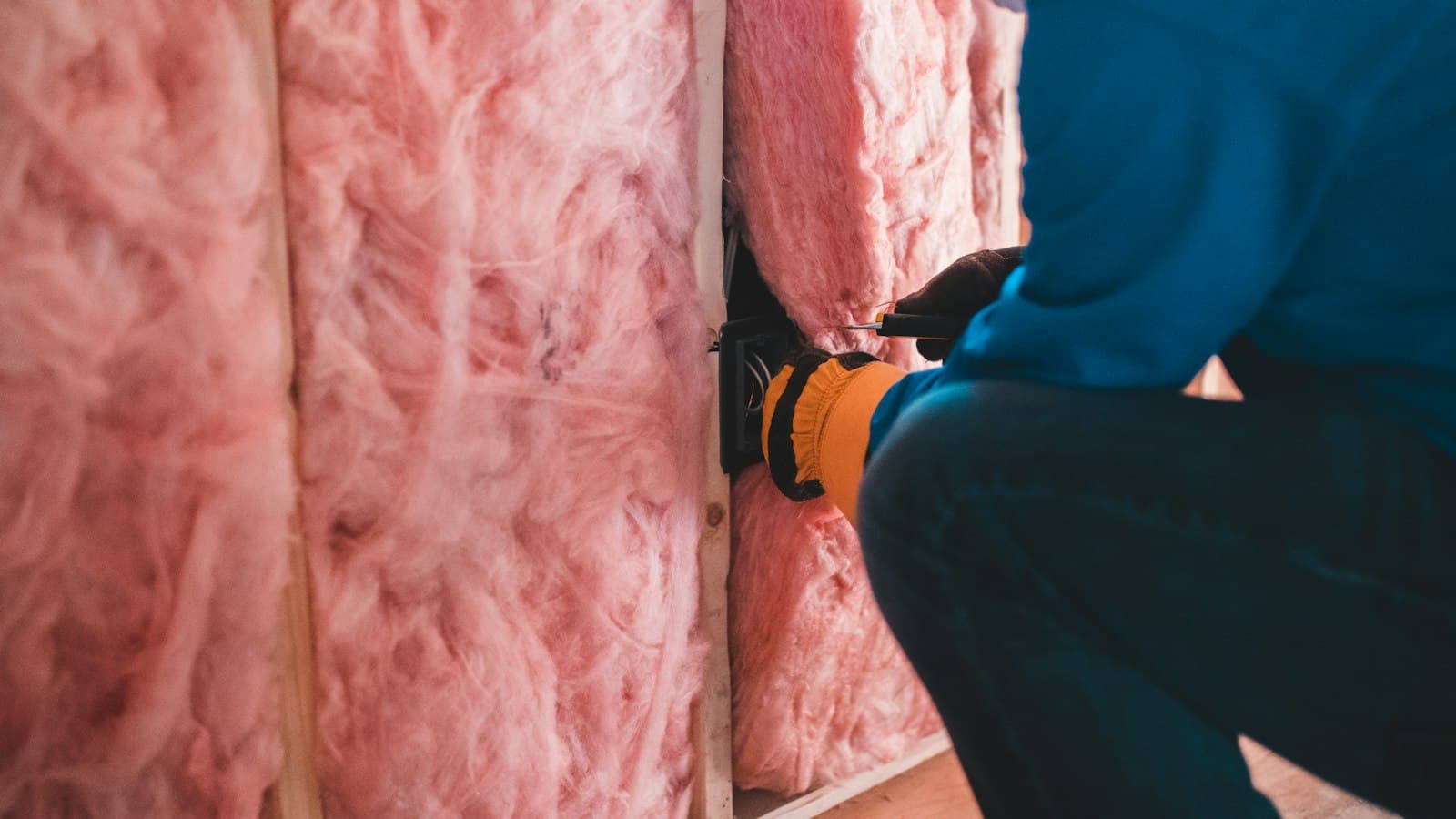 An installer in gloves fitting pink insulation batts into a wall frame