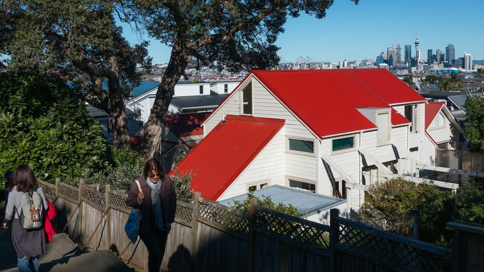 Two people walking up a footpath beside a white house with a red roof, with the Auckland city skyline behind