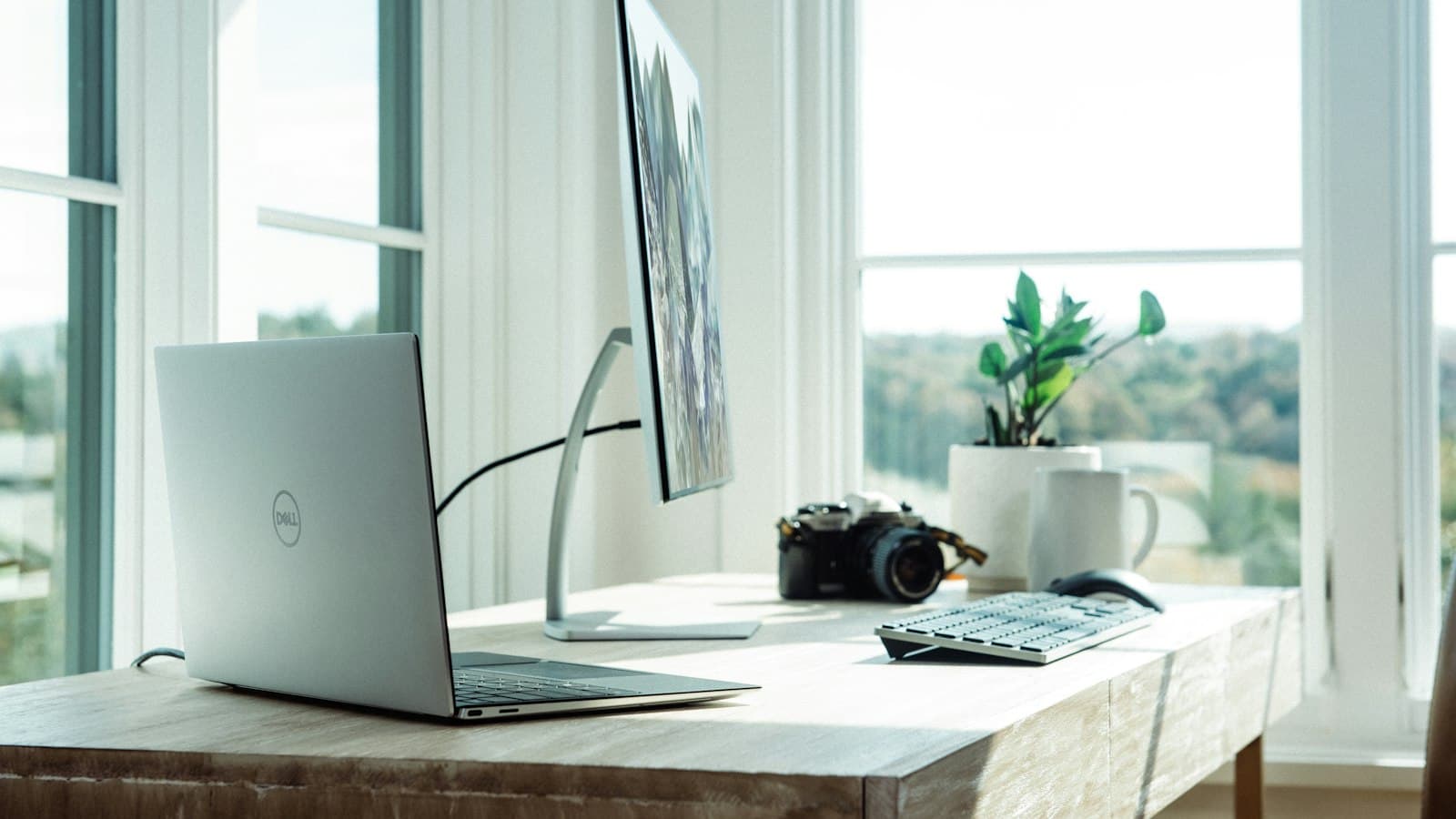 A laptop, monitor, keyboard and camera on a wooden desk beside a large sunny window