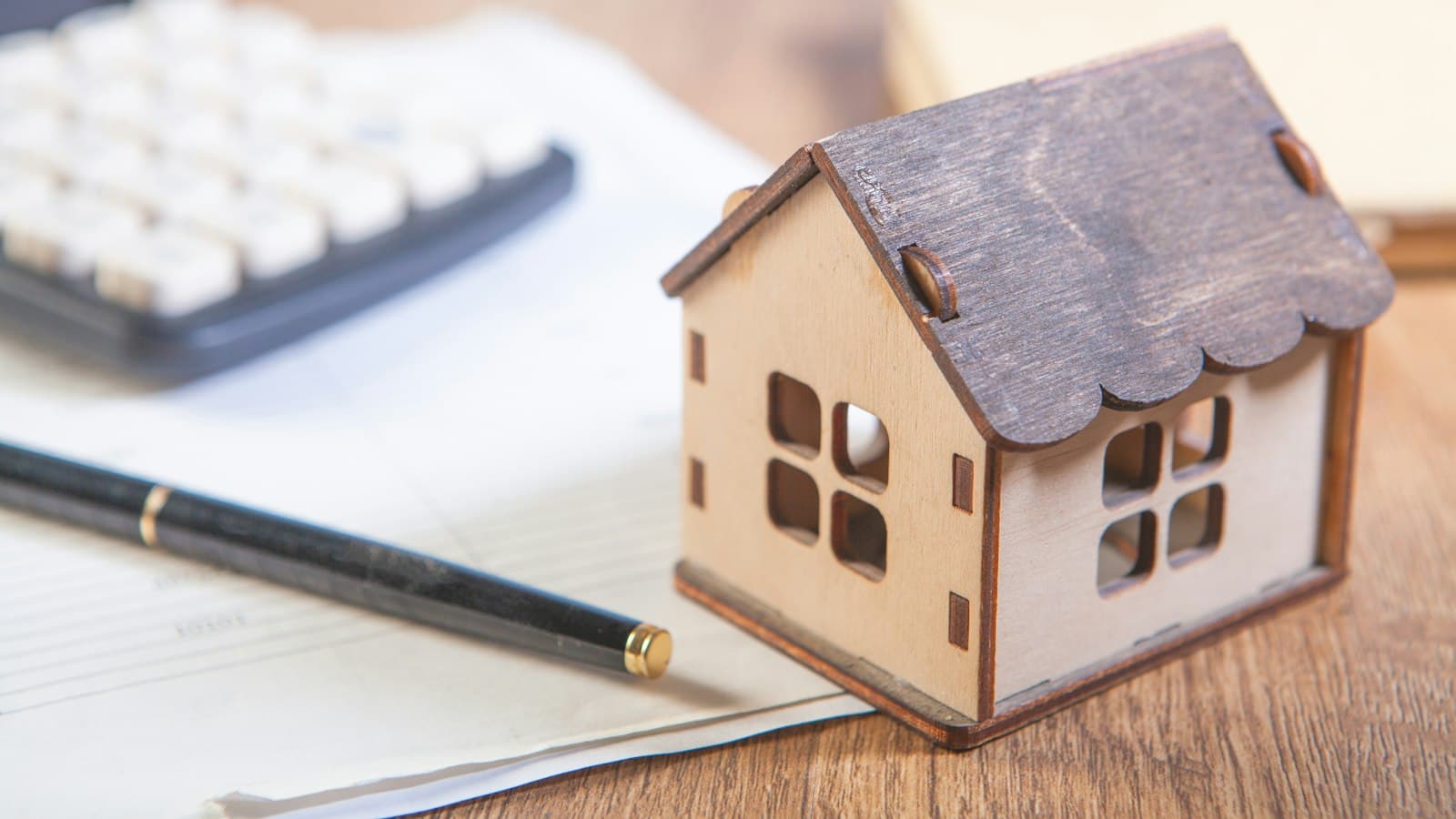 A small wooden model house beside a calculator and pen on a desk