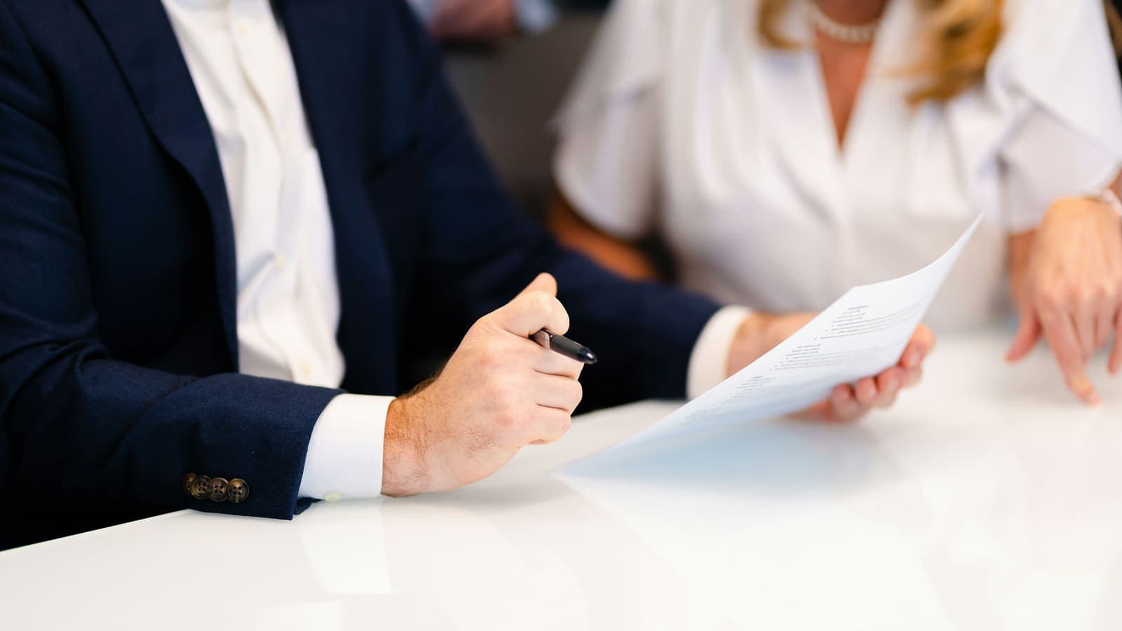 A man in a navy suit holding a pen and a document at a white table, with a woman beside him