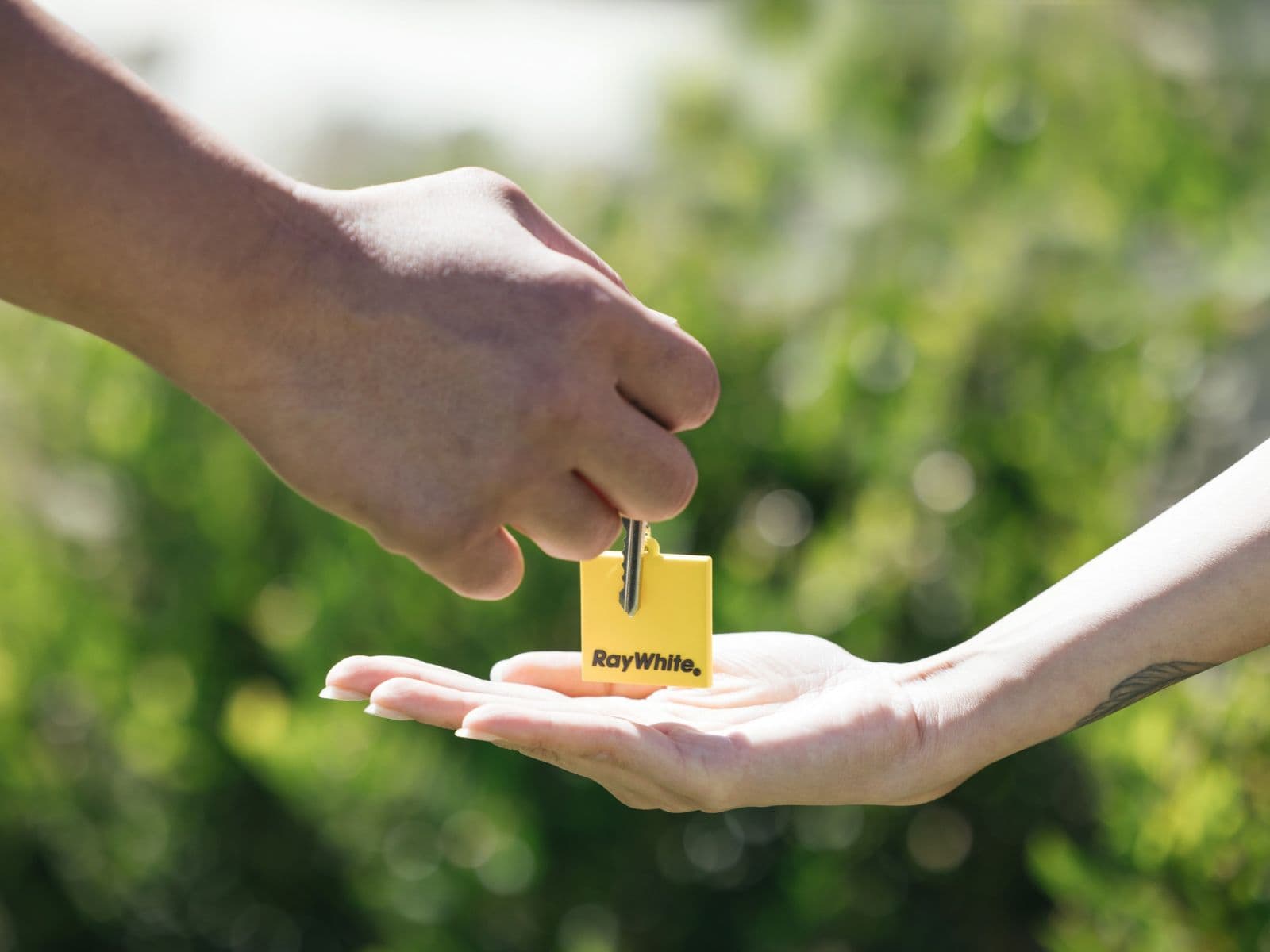 A hand passing a Ray White keyring into an open palm, outdoors against green foliage